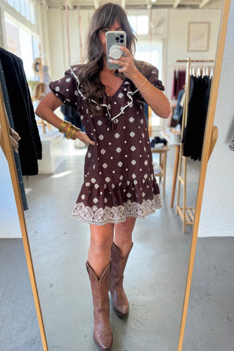 Woman taking a mirror selfie wearing a brown polka dot dress and brown boots in a clothing store.