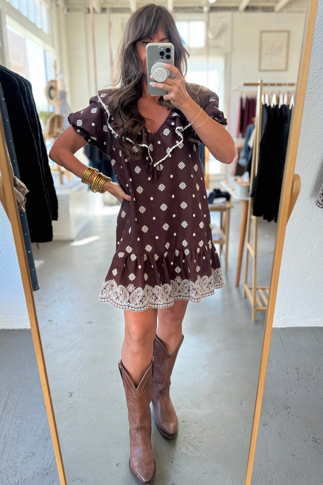 Woman taking a mirror selfie wearing a brown polka dot dress and brown boots in a clothing store.