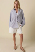 Load image into Gallery viewer, Woman wearing a light blue button-up shirt and white shorts on a beige background