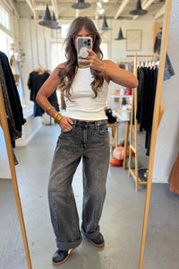 Person taking a mirror selfie wearing a white tank top and gray jeans in a clothing store.