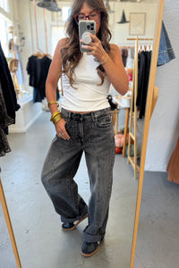 Person taking a mirror selfie wearing a white tank top and dark jeans in a clothing store.