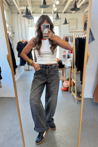 Person taking a mirror selfie wearing a white tank top and gray jeans in a clothing store.