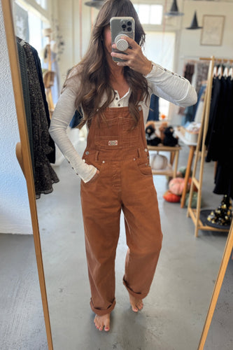 Person wearing brown overalls taking a mirror selfie in a clothing store.