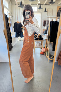 Person wearing brown overalls taking a mirror selfie in a clothing store.