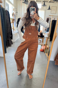 Person wearing brown overalls taking a mirror selfie in a clothing store.
