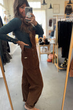 Load image into Gallery viewer, Person wearing a green top and brown jumpsuit taking a mirror selfie in a clothing store.
