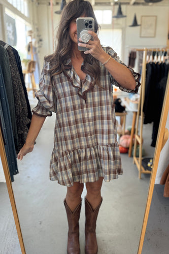 Woman taking a mirror selfie wearing a plaid dress and brown boots in a clothing store.