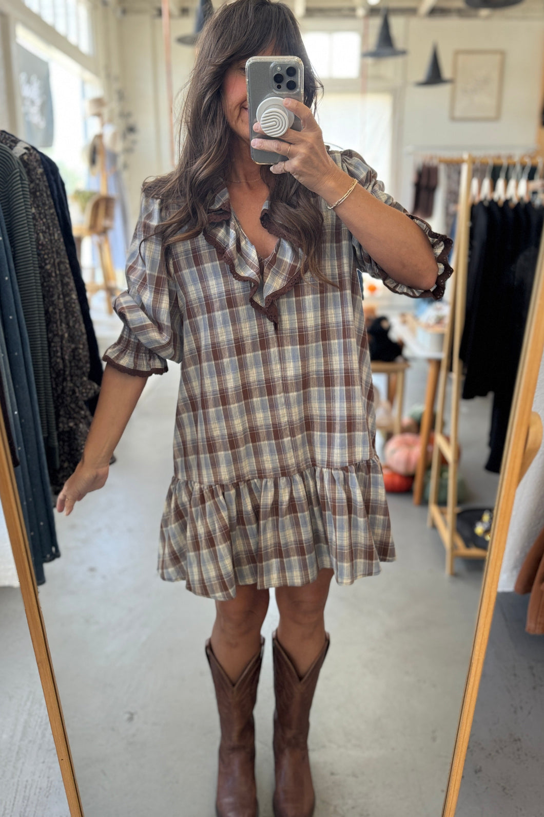 Woman taking a mirror selfie wearing a plaid dress and brown boots in a clothing store.