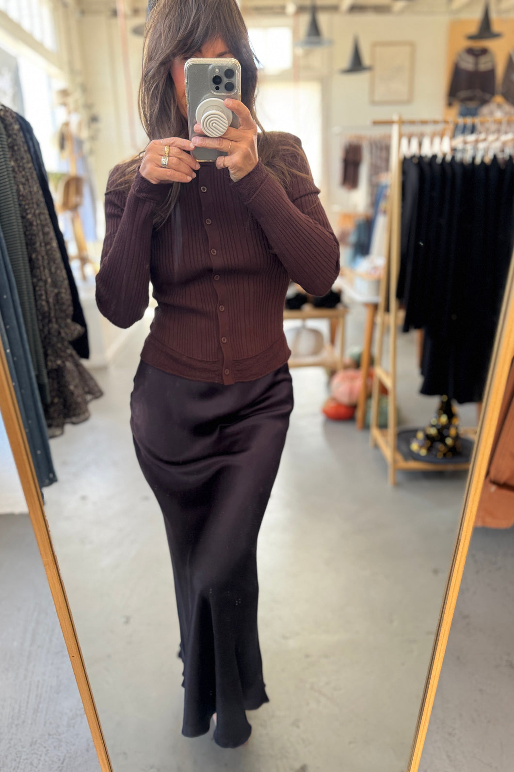 Person taking a mirror selfie wearing a brown cardigan and dark pants in a clothing store.