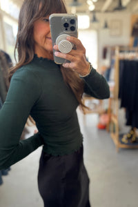 Person taking a mirror selfie wearing a green long-sleeve top and dark pants in a store setting.