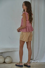Load image into Gallery viewer, Woman wearing a red checkered top and beige shorts standing against a white wall.