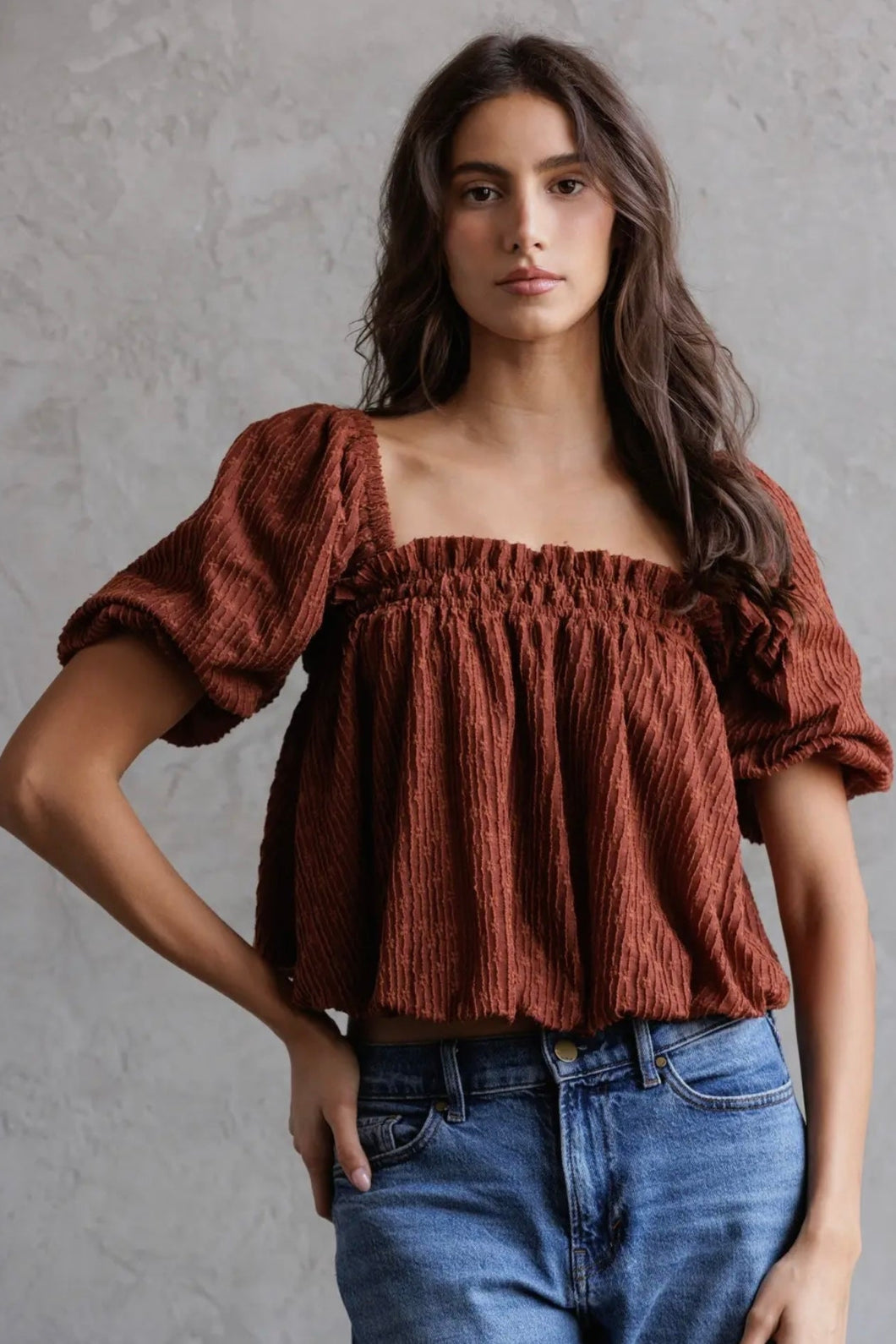Woman wearing a brown top with puffed sleeves and blue jeans against a gray background