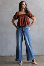 Load image into Gallery viewer, Woman wearing a brown top and blue jeans against a gray wall.