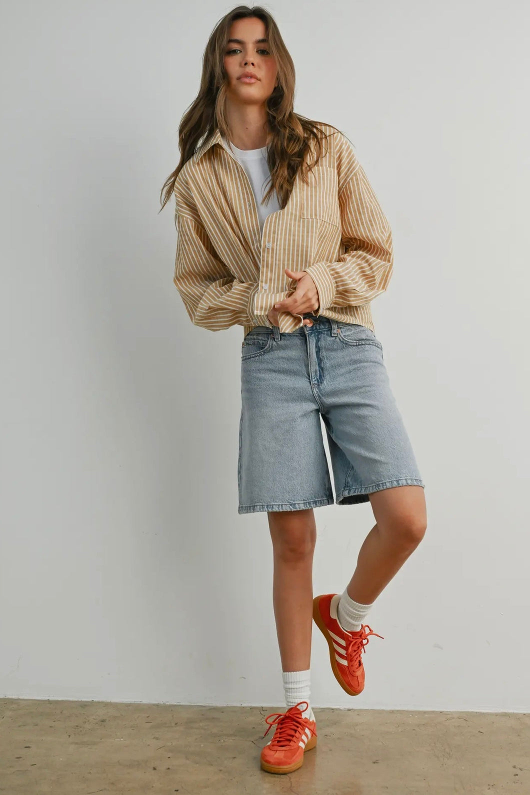 Woman wearing a beige cardigan, white shirt, denim shorts, and red sneakers against a plain background