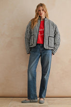 Load image into Gallery viewer, Woman wearing a checkered jacket, red shirt, and blue jeans against a beige wall.