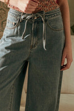 Load image into Gallery viewer, Person wearing light blue jeans with a tie-waist detail, standing indoors.