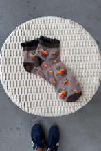 Load image into Gallery viewer, Pair of floral-patterned socks on a woven stool with a person&#39;s feet in blue sneakers.