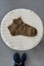 Load image into Gallery viewer, Brown sock on a textured white surface with a person&#39;s foot in blue shoes in the background.