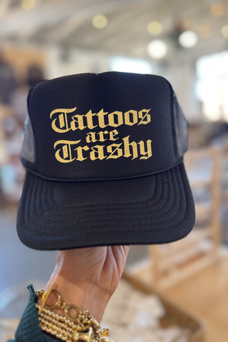 Black cap with 'Tattoos are Trashy' text in gold held by a person.