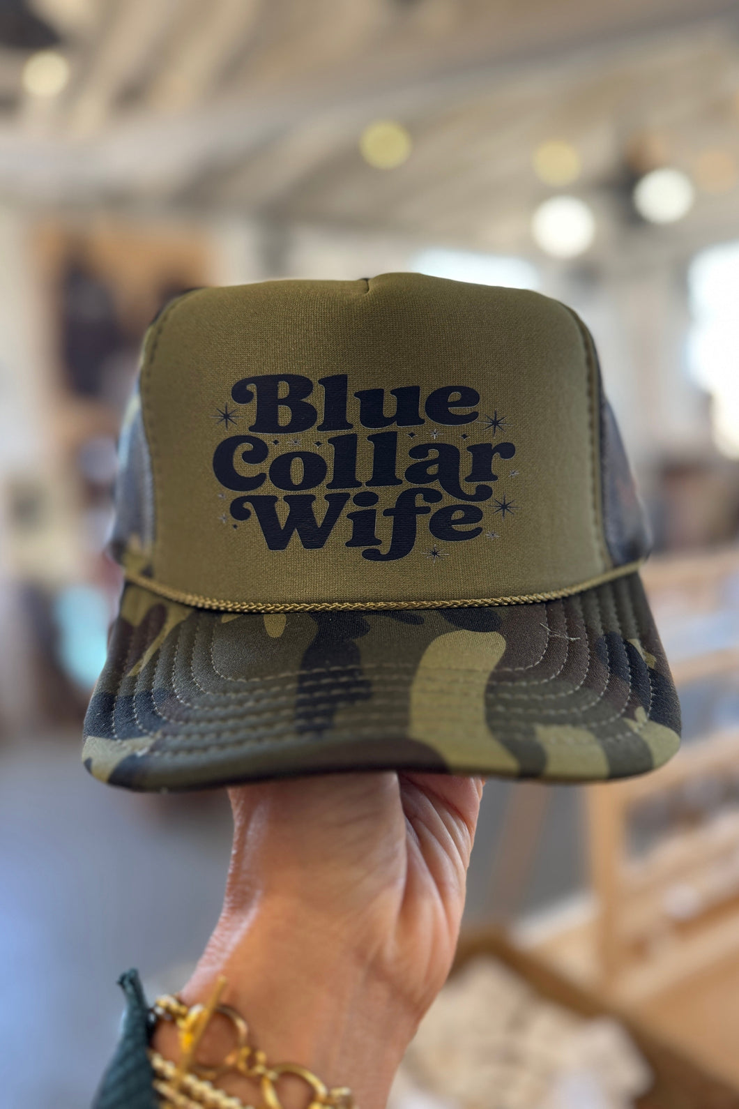 Camouflage cap with 'Blue Collar Wife' text held by a person indoors.