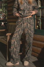 Load image into Gallery viewer, Person wearing a camouflage outfit standing in front of a wooden cabin.