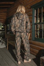Load image into Gallery viewer, Person wearing a camouflage outfit standing on a wooden porch of a log cabin.