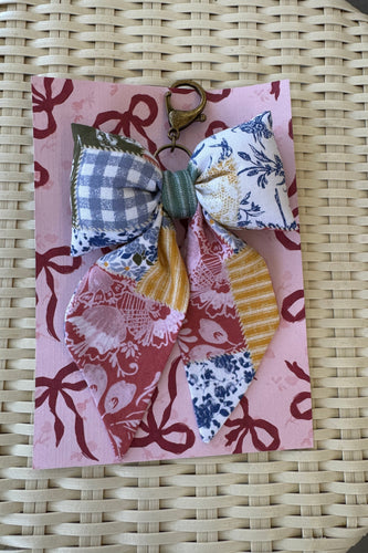 Decorative fabric bow on a pink card with floral and checkered patterns, placed on a woven surface.