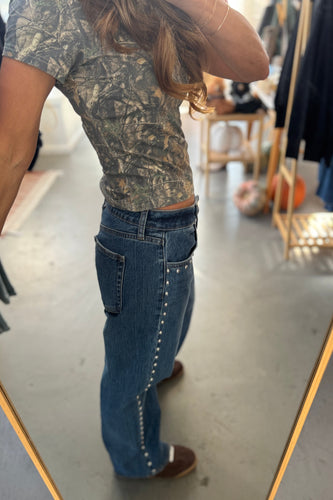 Person wearing a camouflage shirt and blue jeans standing in front of a mirror.