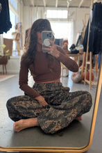 Load image into Gallery viewer, Person sitting on the floor taking a mirror selfie wearing a brown top and camouflage pants.