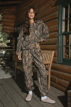 Load image into Gallery viewer, Person wearing a camouflage outfit standing on a wooden porch of a log cabin.