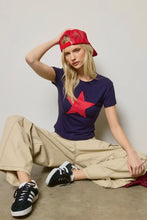 Load image into Gallery viewer, Woman wearing a purple t-shirt with a red star, beige pants, and black sneakers, sitting on the floor.