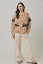Load image into Gallery viewer, Woman wearing a beige knitted cardigan with black swan designs on a plain background