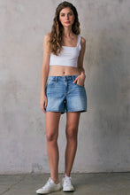 Load image into Gallery viewer, Woman wearing a white tank top and blue denim shorts against a gray concrete wall.