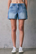 Load image into Gallery viewer, Person wearing light blue denim shorts against a gray concrete wall.