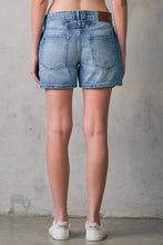 Load image into Gallery viewer, Person wearing light blue denim shorts against a gray concrete wall.