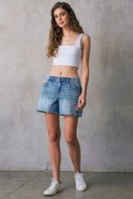 Load image into Gallery viewer, Woman wearing a white crop top and denim shorts against a gray concrete wall.