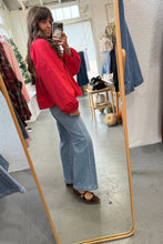 Load image into Gallery viewer, Person taking a mirror selfie wearing a red sweatshirt and blue jeans in a clothing store.