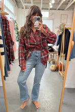 Load image into Gallery viewer, Person wearing a red plaid shirt and blue jeans taking a mirror selfie in a clothing store.