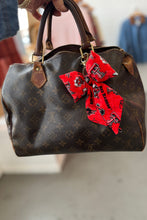 Load image into Gallery viewer, Brown handbag with a red bow attached, held by a person.