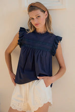 Load image into Gallery viewer, Woman wearing a navy blue top with ruffled sleeves and a white skirt against a beige wall.