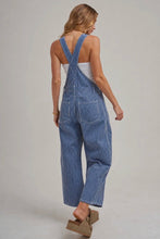 Load image into Gallery viewer, A woman shown from the back wearing blue and white pinstriped denim overalls over a white tank top, paired with brown wedge sandals, against a light gray studio background.