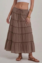 Load image into Gallery viewer, A woman wearing a brown and beige gingham tiered maxi skirt with a drawstring waist, styled with a white crop top and brown strappy sandals against a light gray studio background.