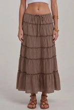 Load image into Gallery viewer, A woman wearing a brown and beige gingham tiered maxi skirt with a drawstring waist, paired with brown strappy sandals and gold bracelets, against a light gray studio background.