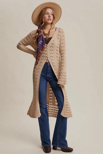 Woman wearing a long beige cardigan, blue jeans, and a hat on a plain background