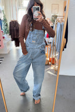 Load image into Gallery viewer, Person wearing light blue denim overalls taking a mirror selfie in a clothing store.