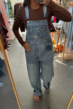 Load image into Gallery viewer, Person wearing blue jeans and a brown jacket standing on a concrete floor with stacks of folded clothes in the background.